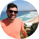 RE4ORM synthetic turf recycling &ndash; A man wearing sunglasses and a coral T-shirt smiles at the camera with Kareela Oval and ocean waves in the background on a sunny day.