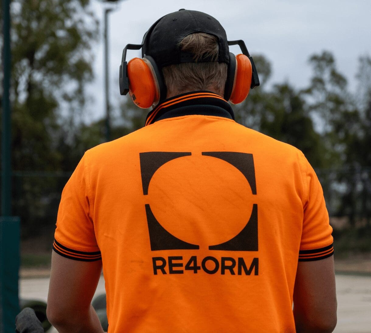 RE4ORM synthetic turf recycling &ndash; A person wearing an orange shirt with "RE4ORM" and a square logo on the back, along with orange ear defenders and a black cap, stands outdoors facing away from the camera.