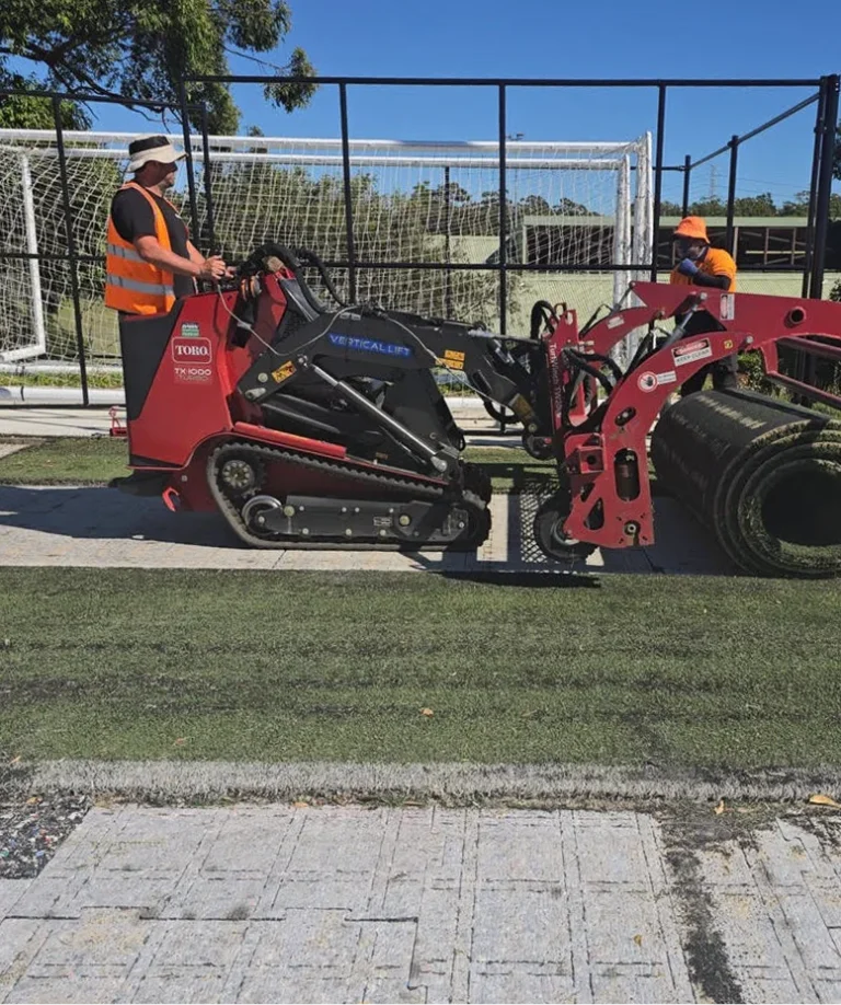 RE4ORM synthetic turf recycling &ndash; Two workers operate a red machine to roll up artificial turf at Kareela Oval, with football goals and green grass visible in the background under a clear blue sky.