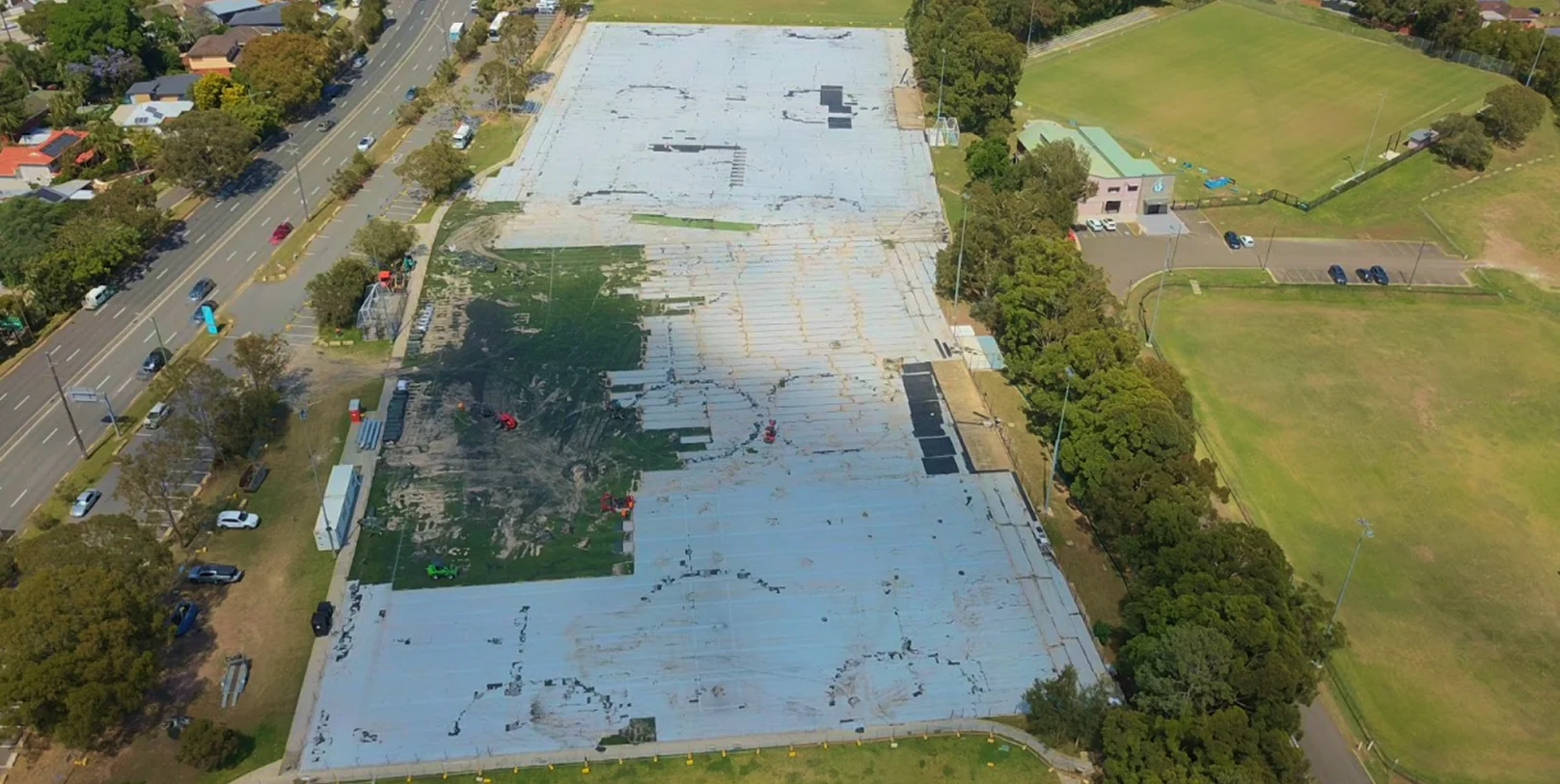 RE4ORM synthetic turf recycling &ndash; Aerial view of a large, mostly empty concrete car park near Kareela Oval, with patches of greenery and equipment, surrounded by trees, grass fields, and a road lined with houses on the left side.