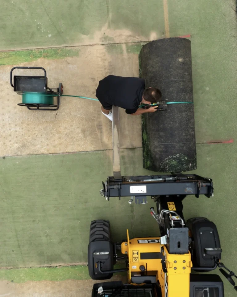 RE4ORM synthetic turf recycling – A person secures a strap around a large rolled-up piece of artificial turf next to a reel at Kensington Banks, while a yellow construction vehicle prepares to lift the roll on a partially tarmacked surface.