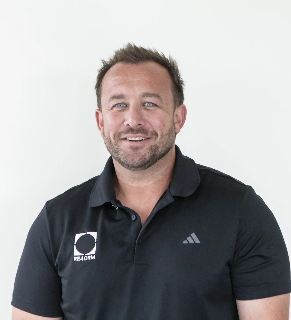 RE4ORM synthetic turf recycling &ndash; A man with short brown hair and a beard smiles at the camera. He is wearing a black collared shirt with an Adidas logo and another circular logo that says "REFORM" on the left side. The background is plain white.