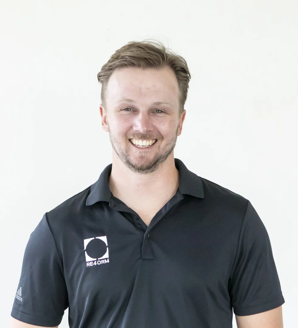 RE4ORM synthetic turf recycling &ndash; A man with short light brown hair and a beard smiles at the camera. He is wearing a dark polo shirt with a RE4ORM logo on the left side. The background is plain and light-coloured.
